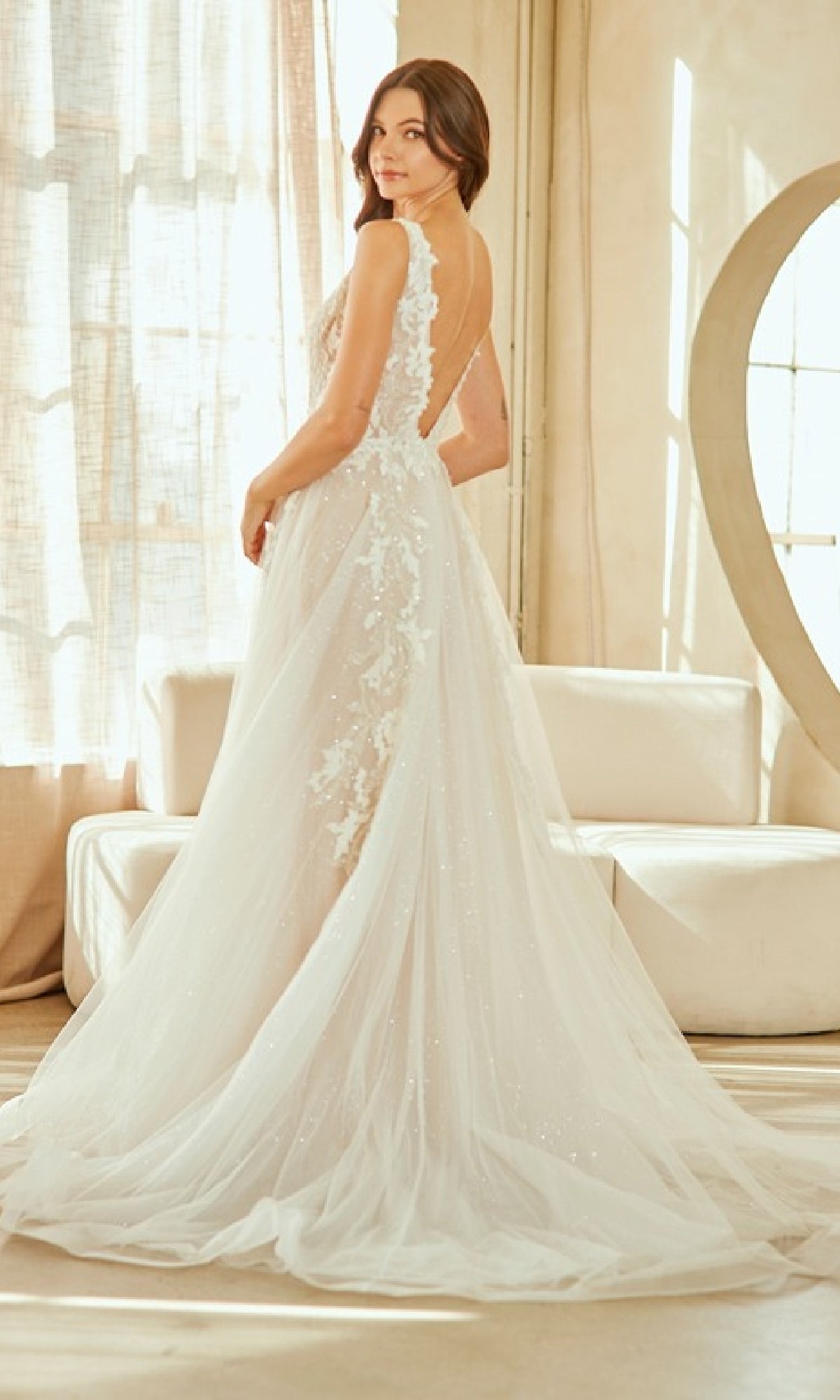 A woman stands indoors in the Amelia Couture 2140, a white, floor-length wedding dress with tulle, lace embroidery, sheer details, and a corset bodice as she faces slightly away from the camera.